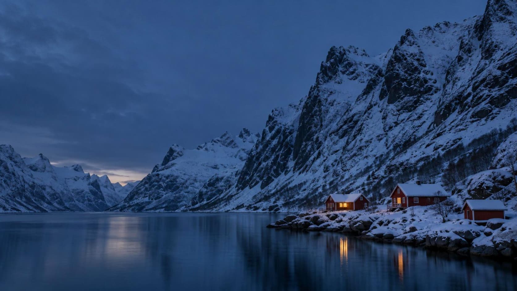 Verschneiter Fjord zur blauen Stunde mit roten Holzhütten am Ufer