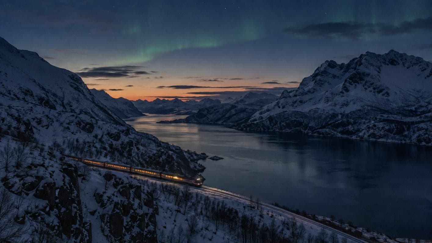 Beleuchteter Zug an einer Bahnstrecke hoch über einem verschneiten Fjord in der Dämmerung