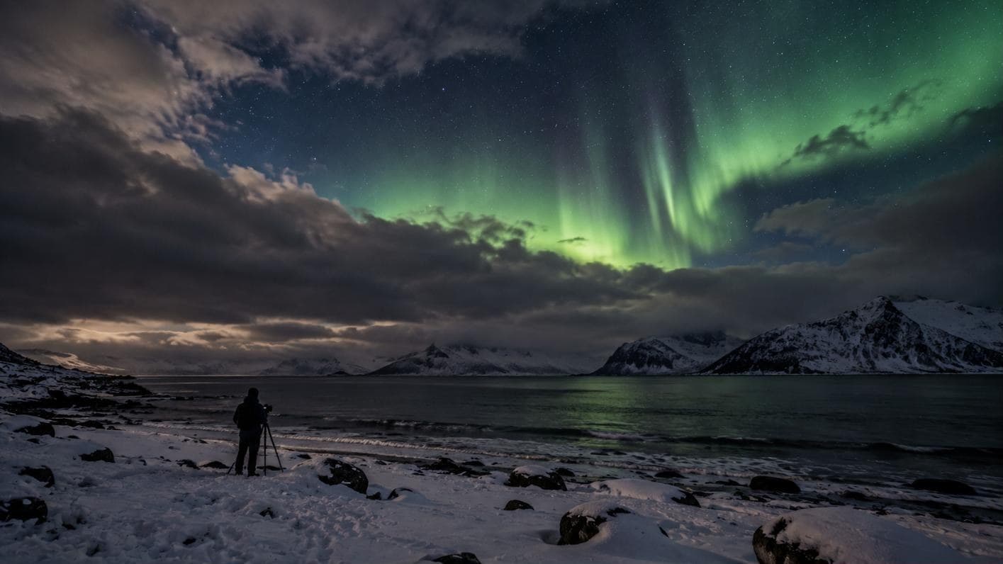 Fotograf mit Stativ am Strand, Nordlicht bricht durch eine Wolkenlücke