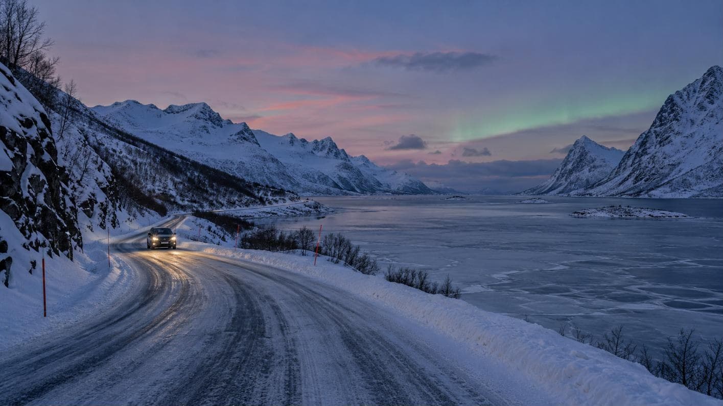 Auto auf verschneiter Küstenstraße am zugefrorenen Fjord zur blauen Stunde mit erstem Nordlicht