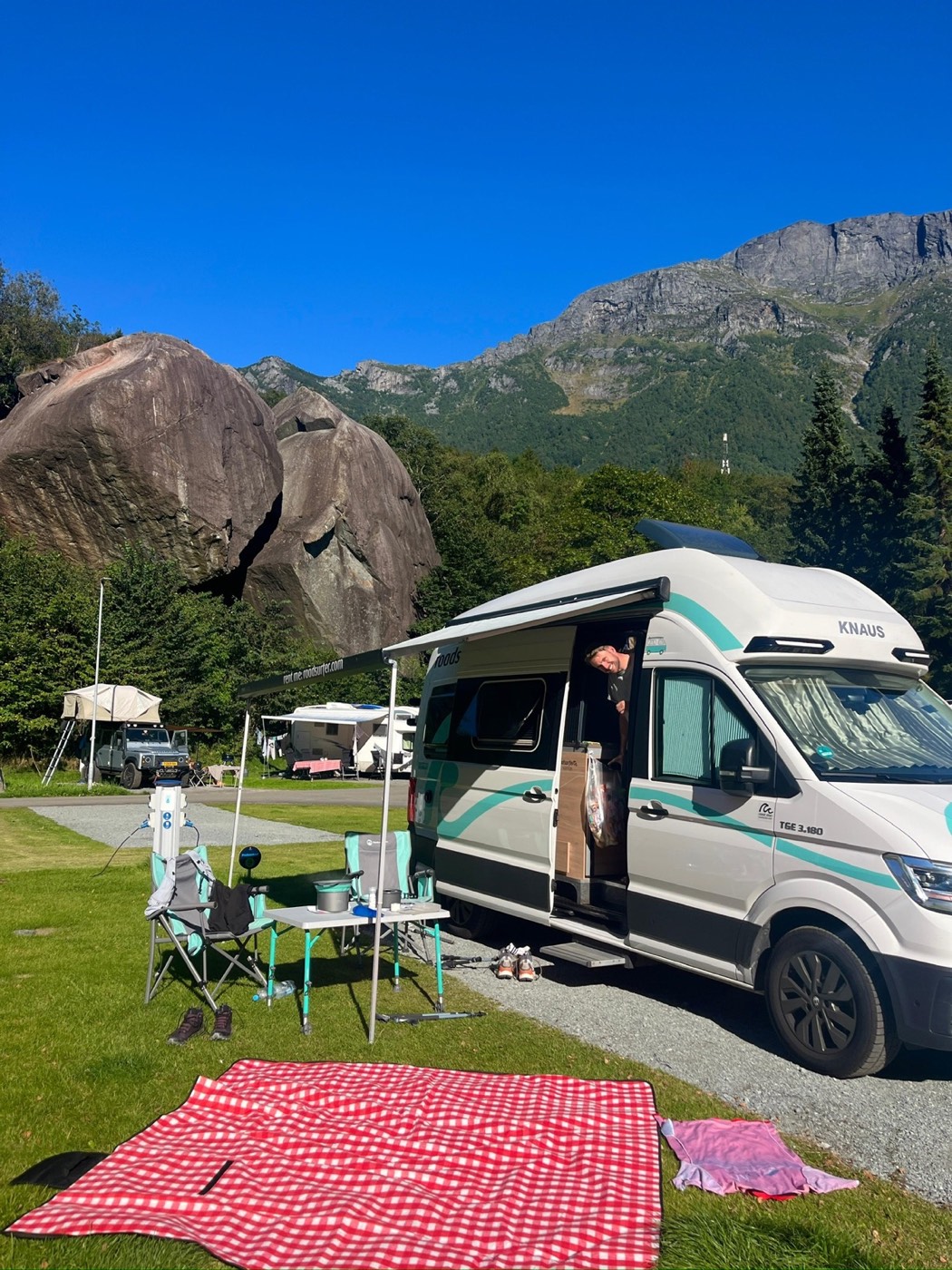 Camper mit ausgefahrener Markise auf einem Campingplatz, dahinter große Felsblöcke und steile Berge